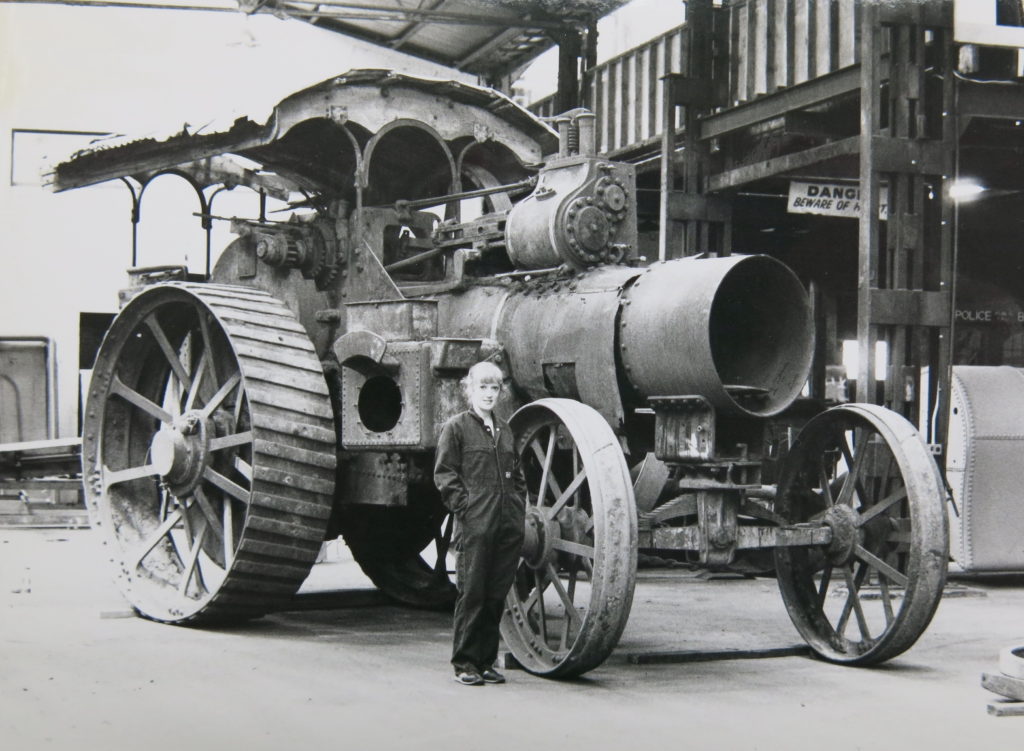 A woman wearing overalls is standing inside an old factory building in front of a large steam traction engine which has a canopy roof. A traction engine is a self-propelling steam road vehicle. The fuel was burnt and steam raised in a locomotive-type boiler. 